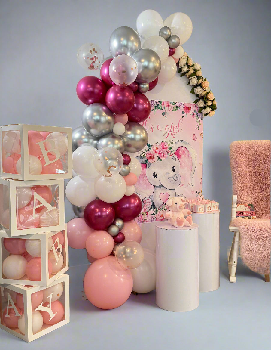 It’s a Girl baby shower setup with pink, white, and silver balloon arch, floral accents, BABY blocks, teddy bear, and themed backdrop