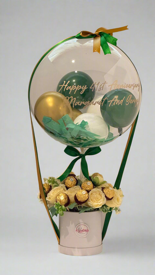 Decorative balloon with balloons, flowers, and chocolates inside a clear dome on a white background.